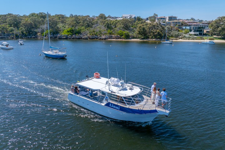 Small boat named Linni with people on deck, sailing in a scenic river with other boats and houses in the background.