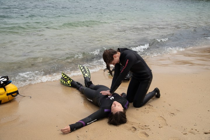Person giving CPR on a beach to another person in wetsuits.