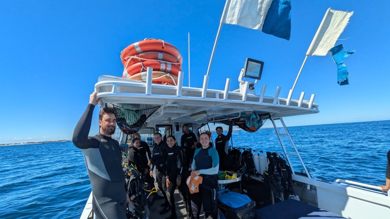 People in wetsuits on a boat with dive gear and flags, under a clear blue sky.
