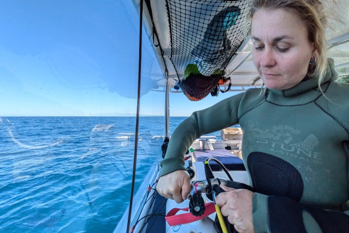 Person in wetsuit adjusting scuba gear on a boat with ocean view.