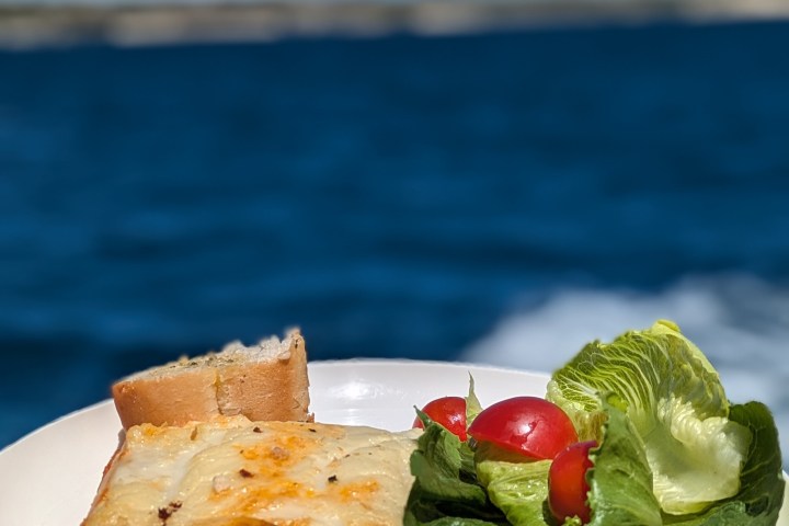 Plate with lasagna, bread, and salad on boat with ocean background.