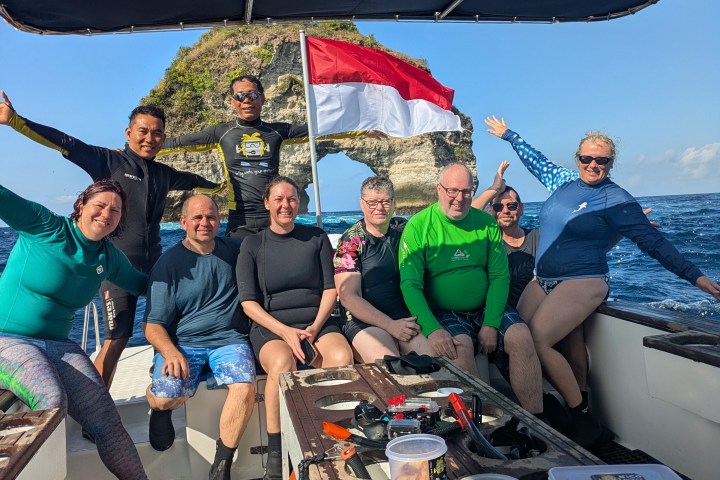 Group posing on a boat with Indonesian flag and rocky arch in the background.