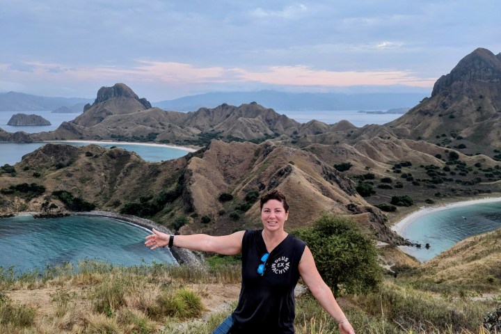 Person posing on a hill with arms outstretched, overlooking a coastal landscape.