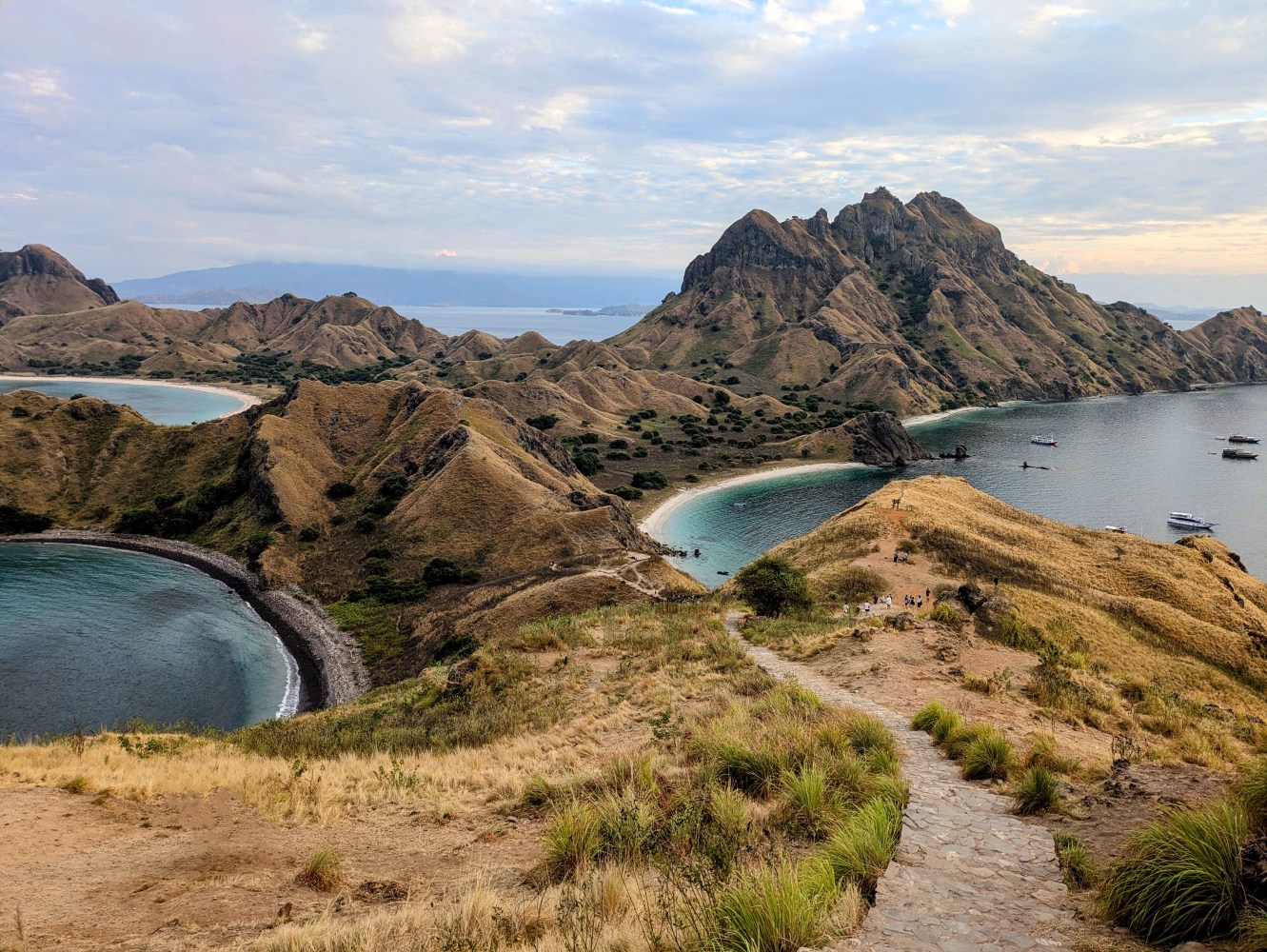 Mountainous island landscape with beaches and boats in the ocean, under a cloudy sky.