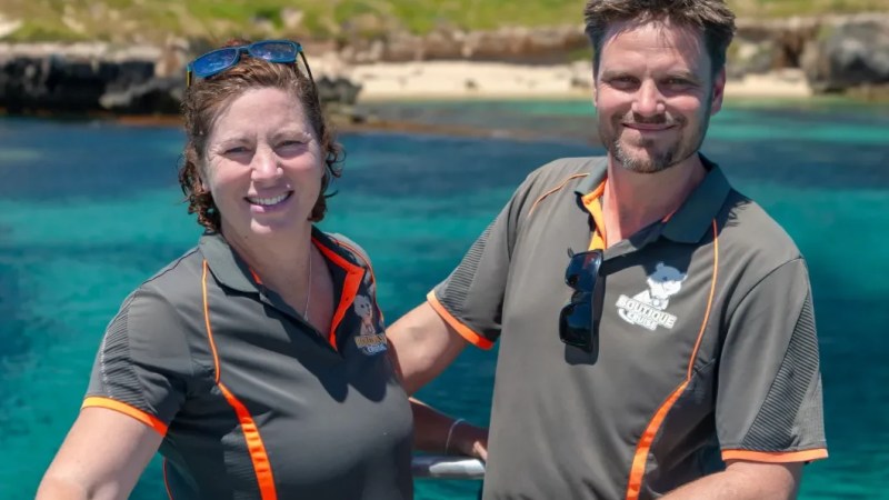 Two people smiling on a boat with clear water and land in the background.