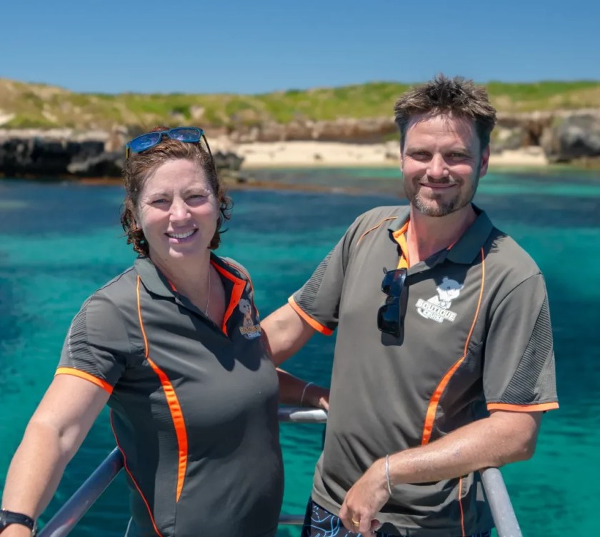 Two people smiling on a boat with clear water and land in the background.
