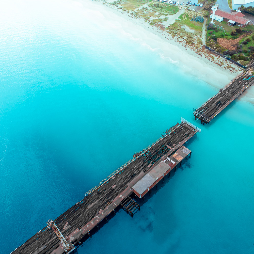 Aerial view of a pier extending into clear blue water, with a sandy shoreline and sparse buildings.